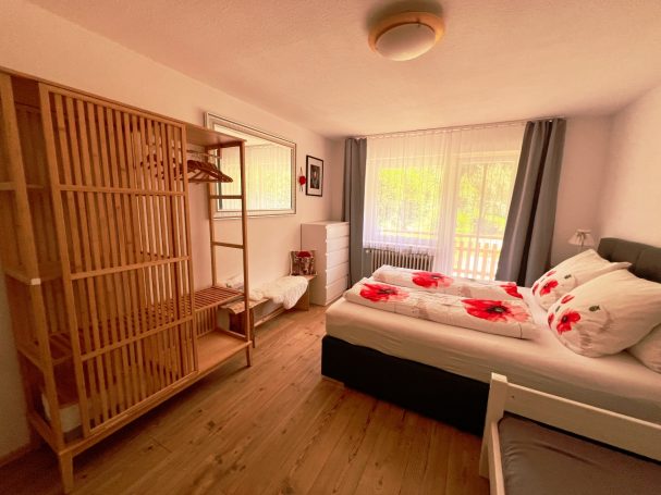 Helles Schlafzimmer mit Bett, Rattanregal und großen Fenstern.