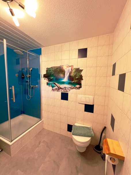 Badezimmer mit Dusche, Toilette und Wandbild eines Wasserfalls.