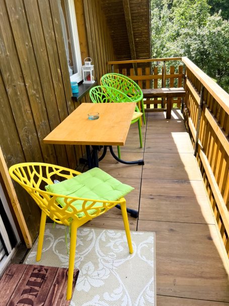Balkon mit Holztisch und bunten Stühlen in einem Waldumfeld.